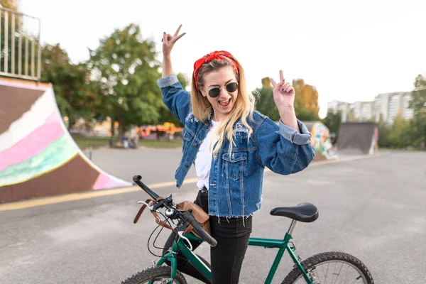 Giovane donna felice sta con una bicicletta sullo sfondo di uno skate park e gioisce con le mani alzate. Gioiosa ragazza hipster in bicicletta in città . — Foto Stock