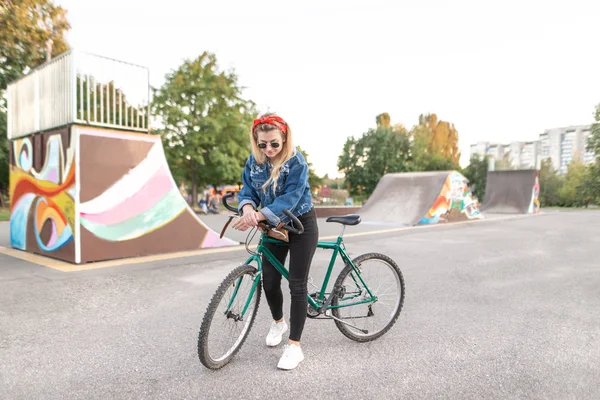 Çekici hipist kız şık giysiler ve güneş gözlüğü ile bir bisiklet bir kaykay arka plan üzerinde duruyor. Şehir içinde Bisiklete binmek bir şık kız portresi. — Stok fotoğraf