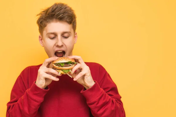 Jovem bonito com uma camisola vermelha segura um hambúrguer em suas mãos, olha e vai comer. Adolescente com fome come um hambúrguer apetitoso em um fundo amarelo. Fast food — Fotografia de Stock