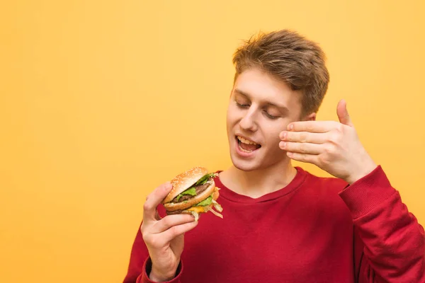 그의 손에 햄버거와 행복한 소년은 신선한 패스트 푸드의 냄새를 흡입하고 노란색 배경에 미소. 청년은 고립된 식욕을 돋우는 햄버거를 냄새맡는다. 패스트푸드 컨셉. — 스톡 사진