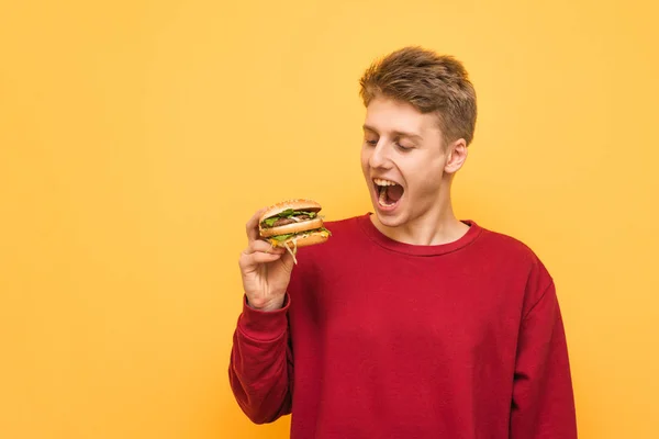 놀란 청년은 노란 배경에 고립된 손에 있는 햄버거를 바라보고 있다. 그의 손에 햄버거와 노란색 배경에 감정적 인 사람의 초상화. 패스트푸드. — 스톡 사진