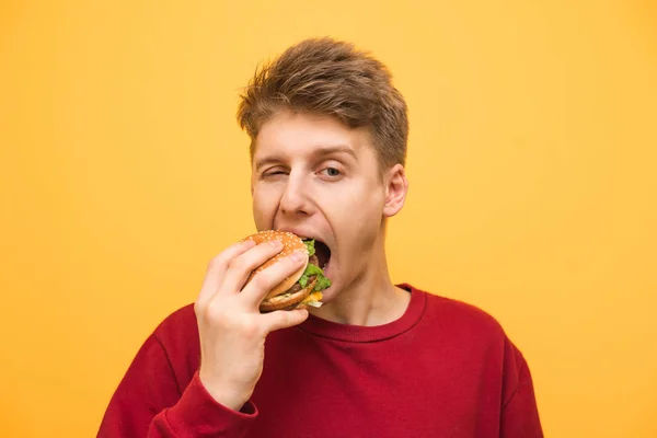 Yakın yukarı bir çocuk portre sarı bir arka planda bir burger yiyor ve kamera bakıyor, Genç adam bir burger ısırır. Fast-Food konsepti. — Stok fotoğraf