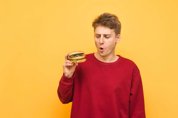 재미있는 젊은 남자는 그의 손에 햄버거를 들고, 노란색 배경에 고립 된 놀란 표정으로 패스트 푸드를 봅니다. 배고픈 십대 와 햄버거 에 그의 손 이다 에 a 노란색 배경. — 스톡 사진