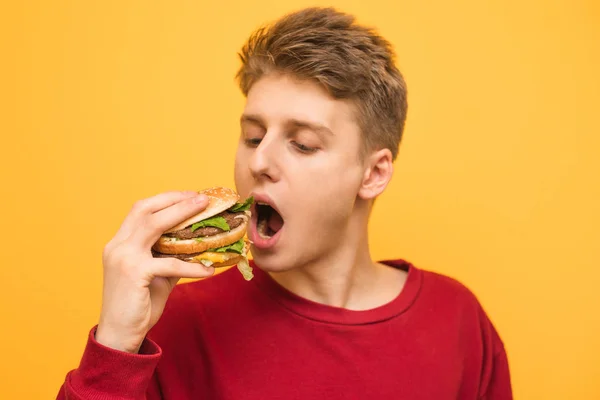 El retrato de cerca de un hombre muerde una hamburguesa apetitosa sobre un fondo amarillo. El tipo come una hamburguesa grande, aislado sobre un fondo amarillo. Concepto de comida rápida . — Foto de Stock