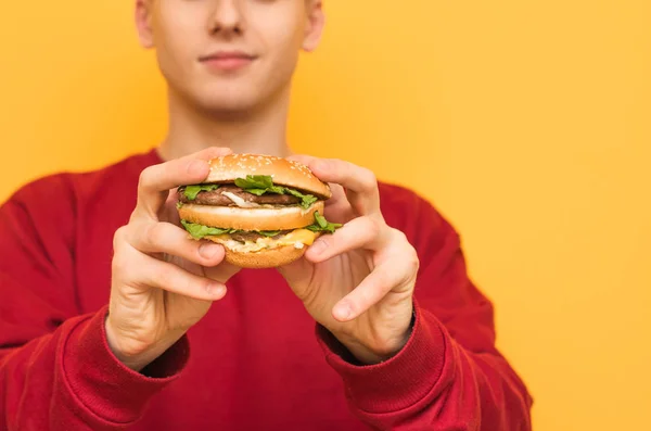写真を閉じます。男は黄色の背景においしい大きなハンバーガーを手に持っています。ハンバーガーに集中しろファーストフードの男、孤立した黄色。コピースペース. — ストック写真