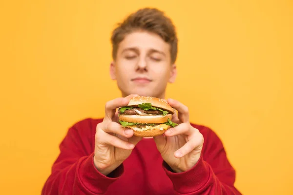 눈을 감은 잘생긴 남자는 카메라에 맛있는 큰 햄버거를 보여줍니다, 노란색 배경에 고립 된 그의 손에 들고. 젊은 남자의 손에 식욕을 돋우는 햄버거에 초점을 맞춥니다. 패스트푸드 — 스톡 사진