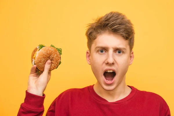 노란 배경에 햄버거를 들고 배고픈 표현 소년의 초상화는 노란색 배경에 카메라에 놀랐다. 그의 손에 햄버거와 감정적 인 젊은 남자. 격리 — 스톡 사진