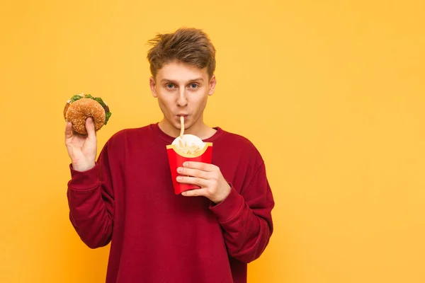 Joven guapo con ropa casual come papas fritas y tiene una hamburguesa en sus manos, mira a la cámara, aislado sobre un fondo amarillo. Estudiante hambriento come comida rápida . — Foto de Stock