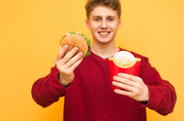 Happy student in Casual kleding houdt een hamburger en frietjes in zijn handen, glimlacht en kijkt naar de camera op een gele achtergrond. Man met slecht voedsel op een gekleurde achtergrond, geïsoleerd — Stockfoto