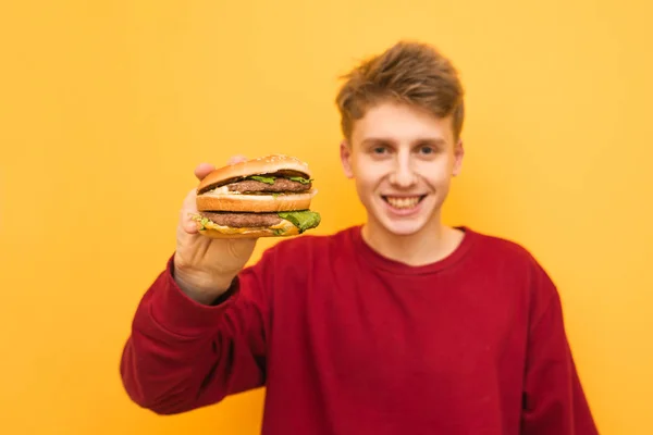 캐주얼 한 옷을 입은 행복한 남자는 그의 손에 식욕을 돋우는 햄버거를 들고 노란색 배경에 고립 된 미소를 지으며 카메라에 보여줍니다. 젊은 남자 쇼 a 좋은 맛있는 햄버거 과 보기 에 이 카메라. — 스톡 사진