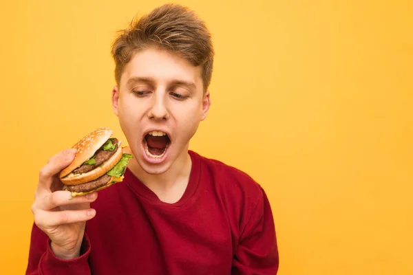 Retrato de um jovem emotivo com um hambúrguer nas mãos sobre um fundo amarelo. Um tipo com fome e emoção segura um hambúrguer nas mãos e grita, isolado. Estudante com muita fome. Espaço de cópia — Fotografia de Stock