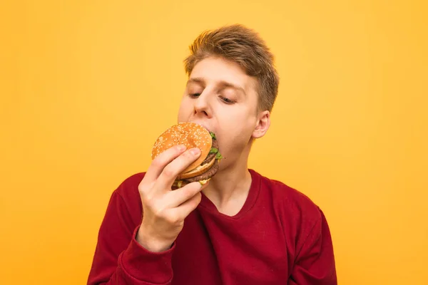 Portret głodnego faceta w odzieży casual gryzie apetyczny Burger na żółtym tle. Młody człowiek zjada Burger, izolowane. Miejsce — Zdjęcie stockowe