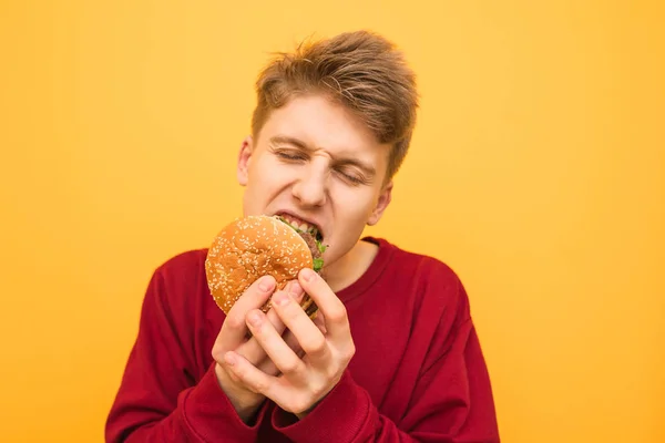 Foto de close-up de um hambúrguer muito faminto jovem mordendo com os olhos fechados em um fundo amarelo. Estudante faminto come comida rápida, close up retrato. Espaço de cópia — Fotografia de Stock