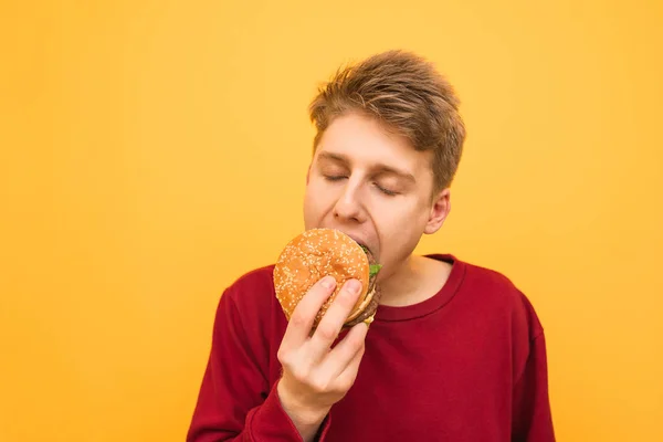 Retrato de close-up de um jovem em roupas casuais morde um hambúrguer com os olhos fechados e tem prazer em um fundo amarelo. O tipo come um hambúrguer apetitoso, isolado. . — Fotografia de Stock