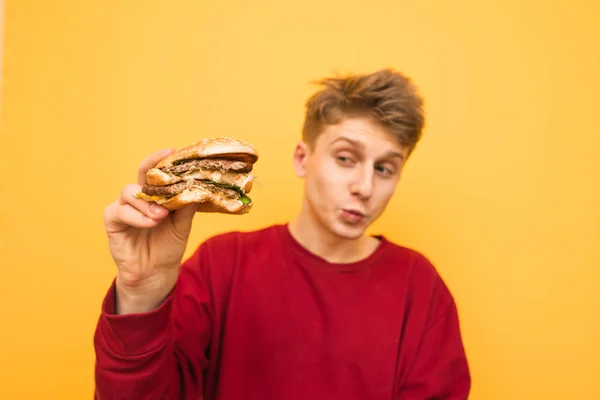 Guapo joven en la ropa casual sostiene una hamburguesa supurante — Foto de Stock