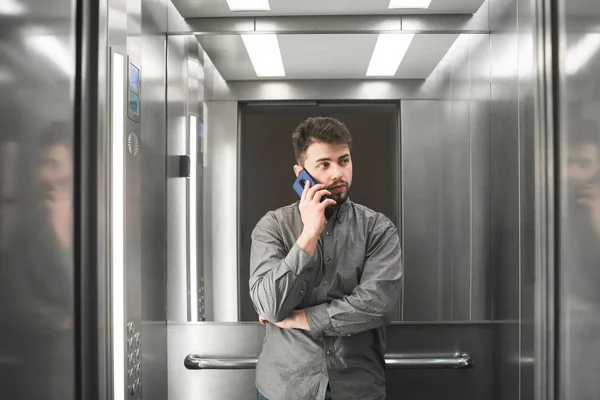 Office worker stands in the elevator, calls on the smartphone and looks away. Business man in a shirt speaks by telephone in an office elevator