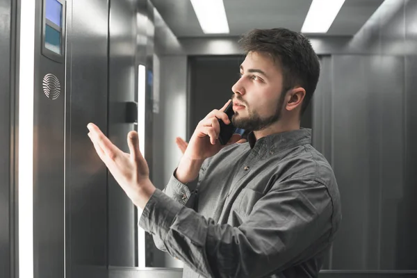 Una empresaria seria explicando algo por teléfono en el ascensor con camisa gris. Empleado barbudo hombre está hablando pacíficamente en su teléfono inteligente y gestos activos — Foto de Stock