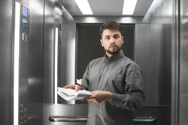Hombre de negocios serio con barba lleva una camisa gris, se para en el ascensor con documentos en las manos y se centra en mirar a la cámara. Retrato de un oficinista en el ascensor de la oficina — Foto de Stock
