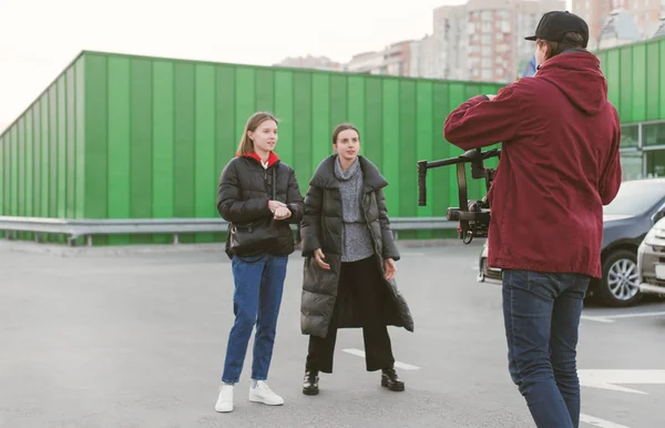 Kamerzysta z stabilizatora w ręku i dwa stylowe dziewczyny strzelać wideo na parkingu. Cameraman bierze wideo z dwóch modeli w ciepłych ubrań. Tworzenie zawartości wideo. Backstage — Zdjęcie stockowe
