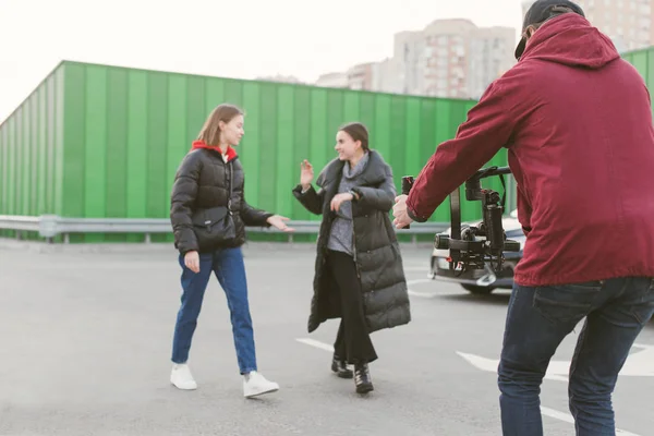 비디오 그래퍼는 안정기로 카메라에 비디오를 찍고, 두 소녀는 거리를 따라 걷고 이야기합니다. 카메라맨은 전문 카메라로 블로거를 위한 비디오를 촬영합니다. 비디오 만들기. 무대 — 스톡 사진