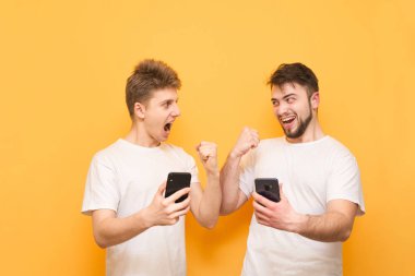 İki neşeli erkek elinde akıllı telefonlar tutun ve sarı bir arka planda izole, birbirlerine bakmak. Sarı bir arka planda mutlu insanlar ellerinde akıllı telefonlar ile sevinirler.