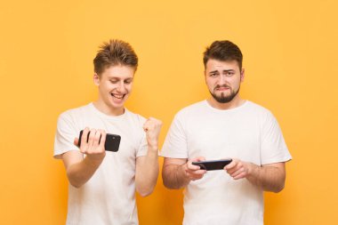 Elinde akıllı telefonlar ile iki genç adam sarı bir arka planda mobil oyunlar oynar. Kazanan ve kaybeden, iki arkadaş mobil oyunlar oynarken portresi. Boşaltmak. Oyun konsepti.