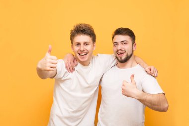 Sarı arka plan üzerinde izole başparmak yukarı gösteren iki mutlu genç erkek portre. Beyaz t-shirt iki mutlu arkadaşlar, kamera bakarak, erkek tarafından sarıldım gülümseyen ve bir başparmak gösteren