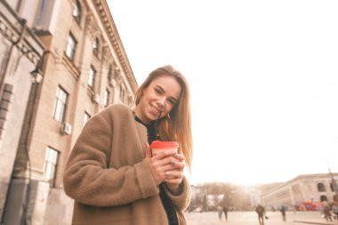 Mutlu bir kızın elinde kahve bir fincan ile sokakta yürürken portre, bir ceket giyiyor, gülümseyen ve gün batımı arka planda kamera bakıyor. Güzel Bayan üzerinde bir yürüyüş