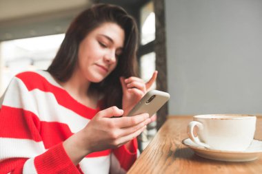 Genç bir kızın portresi yakın yukarı bir kafede bir akıllı telefon kullanır, ekrana bakıyor. Akıllı telefona odaklanın. Kız ellerini yakın yukarıya telefon tutar.
