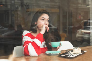 Düşünceli kız elinde bir fincan kahve ve pasta bir tabak ile bir kafede masada yalnız oturur, gün kapalı istirahat ve yan bakıyor