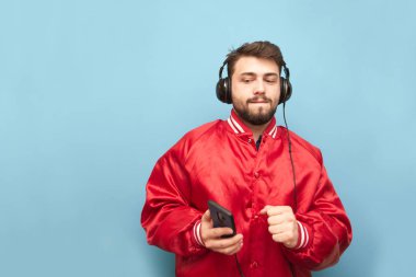 Kırmızı bir ceket ve elinde bir smartphone komik adam mavi bir arka planda kulaklıklar müzik dinlerken. Elinde bir smartphone ile dans kulaklıklar sakallı adam, mavi duvar.