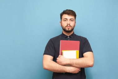 Sakallı bir öğrencinin portresi, karanlık bir gömlek giymiş, elinde kitap ve Notebooklar ile mavi bir arka planda ayakta ve ciddi bir yüz ile kameraya bakıyor. Izole.