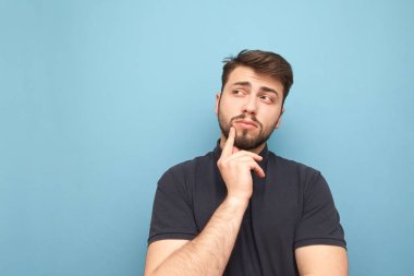 Sakallı ve karanlık bir tişört giymiş adam mavi bir arka planda düşünüyor ve opyspace üzerinde yan görünüyor. Düşünceli yetişkin adam mavi bir arka planda izole edilir.