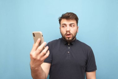 Bir sakal ile hayran adam elinde bir akıllı telefon ile standları ve karışık bir yüz ile karıştı görünüyor. Komik adam elinde bir akıllı telefon tutar, mavi bir arka planda izole