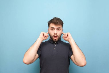 Sürpriz bir yüz ile amotional adam parmaklarıyla kulakları kapatır ve mavi bir arka planda izole kamera, bakıyor. Adam kulakları kapatır, o duymak istemiyor
