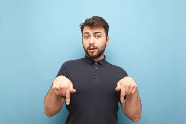 Adam taklit üzerine el aşağı gösterir. Mavi bir arka planda sakal ayakta olan etkileyici adam, kamera bakıyor yüz, boş yere parmaklarını gösteren, koyu bir T-shirt giyiyor