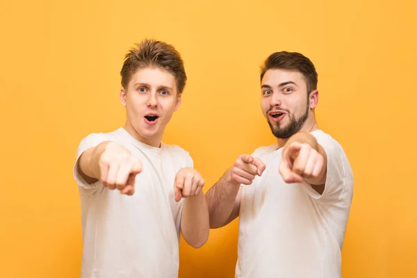 Dos hombres sorprendidos están sobre un fondo amarillo, mirando y mostrando un dedo en la cámara. Dos amigos emocionales llevan una camiseta blanca, aislada sobre un fondo amarillo . — Foto de Stock