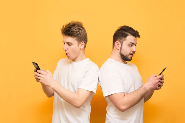 Two surprised men are back-to-back on a yellow background, use a smartphone and look at the screen. Two friends wearing a white T-shirt use a smartphone, isolated on a yellow background