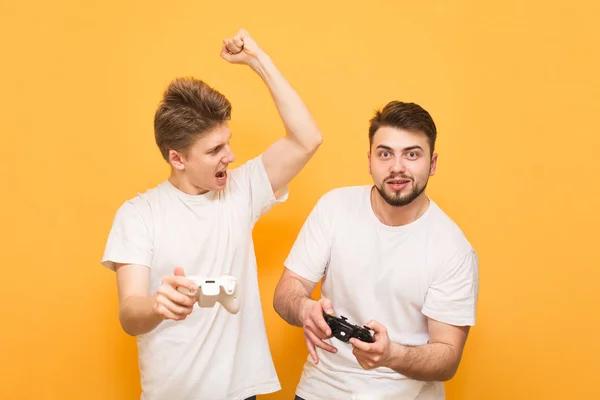 Two men with joysticks in their hands are on a yellow background and play games on the console. Man with a beard focussed looking at the camera, the teenager is angry. Friends are playing games