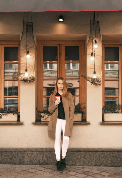 Ritratto di strada di una ragazza elegante in pieno svolgimento, indossando abiti primaverili, in piedi con una tazza di caffè in mano e guardando nella fotocamera. Donna in cappotto beige sullo sfondo di una parete marrone — Foto Stock