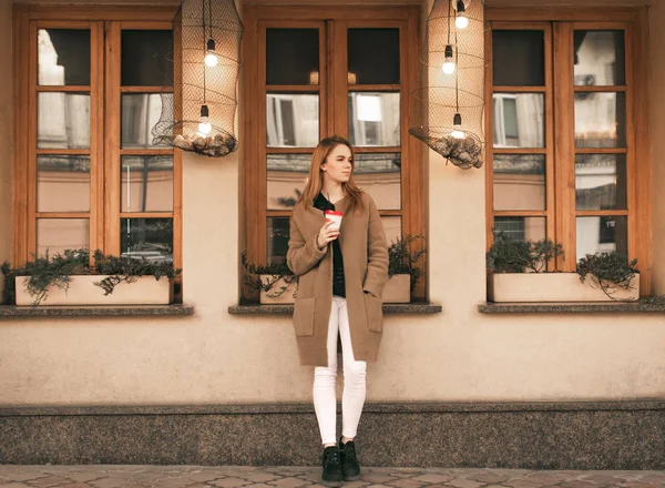 Retrato de uma senhora elegante de comprimento total, de pé com uma xícara de café em suas mãos, vestindo um casaco contra o fundo da parede do café, olhando para o lado. Menina no fundo de uma parede marrom — Fotografia de Stock