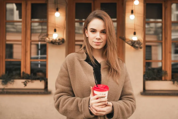 Menina séria fica na rua no fundo da arquitetura, veste um casaco bege, segura uma xícara de café em suas mãos, olha para a câmera e sorri. Retrato de uma mulher elegante — Fotografia de Stock