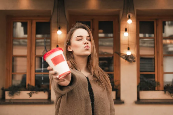 Belle fille debout dans la rue et montrant une tasse de café, baisers avec les yeux fermés.Portrait d'une jolie fille montre un verre de papier de café sur le fond d'un mur de restaurant brun — Photo