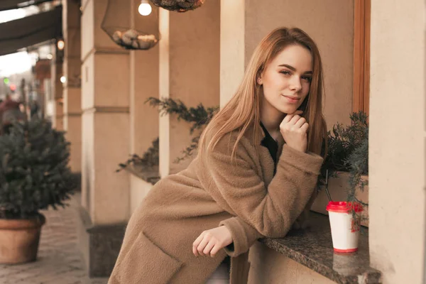 Retrato de rua de uma jovem vestindo um casaco, de pé no fundo de uma parede bege no peitoril da janela de um restaurante com uma xícara de café. Foto de rua de uma menina elegante em roupas de primavera — Fotografia de Stock