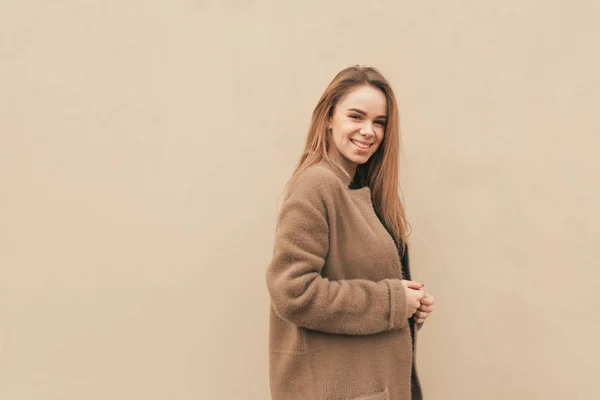 Menina feliz atraente em um casaco fica no fundo de uma parede bege, olha para a câmera e sorri. Isolado. Espaço de cópia — Fotografia de Stock