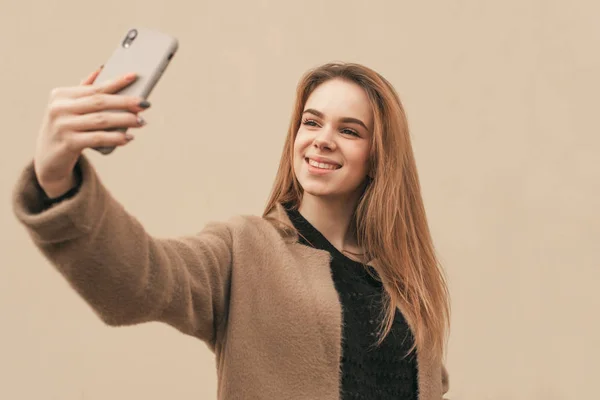 Стильная счастливая девушка носит пальто, стоит на фоне бежевой стены и делает фото на смартфоне, селфи. Модная девушка в весенней одежде делает селфи на фоне стены . — стоковое фото