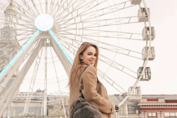 Menina feliz está de pé na rua, no fundo de uma paisagem da cidade, vestindo roupas quentes e uma mochila, olhando câmera e sorrindo. Menina sorridente contra o fundo da roda gigante . — Fotografia de Stock