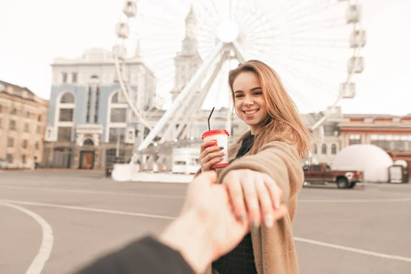 Man holds a happy girl by the hand against a background of urban landscape.Smiling girl with a cup of coffee holds her husband's hand and looks at the camera at the background of the ferris wheel — Stock Photo, Image
