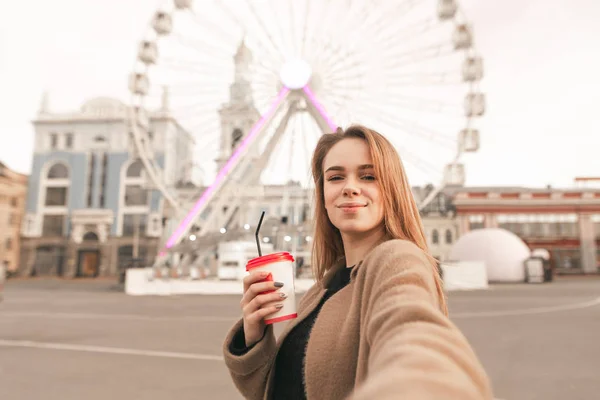 Cute girl in spring clothes, wearing a coat, holding a cup of coffee in her hand and takes selfie on the street background. Attractive lady takes selfie against the background of the ferris wheel