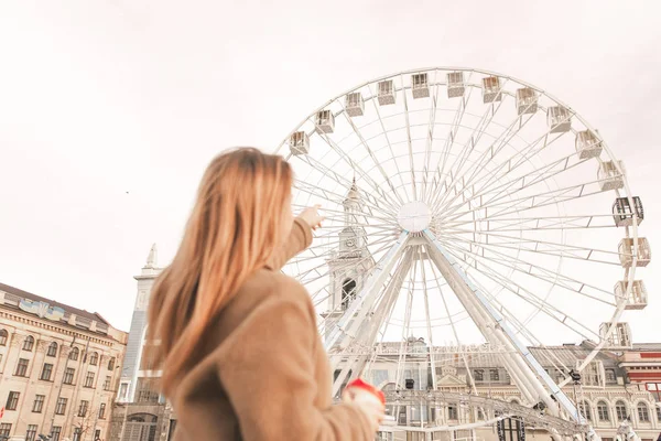 Le dos d'une fille élégante se tient dans la rue avec une tasse de café, portant une tenue décontractée de printemps, montre un doigt sur la roue ferris. Focus sur la roue ferris — Photo